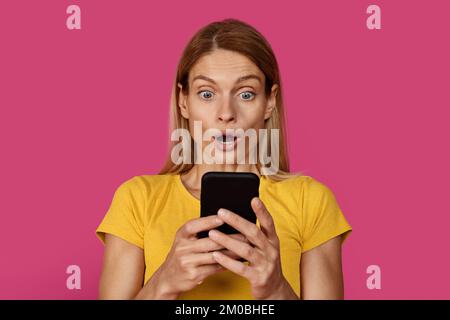 Content surpris mature caucasien blonde femelle dans le t-shirt jaune avec la bouche ouverte regarde le smartphone Banque D'Images