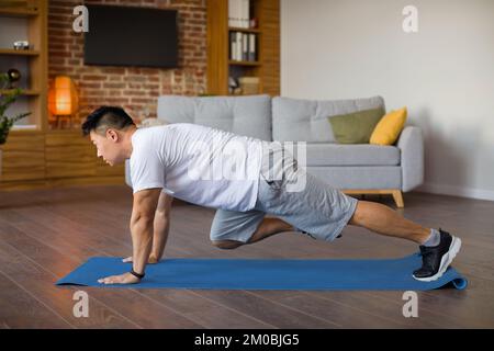 Homme asiatique mature actif faisant des exercices de alpinistes de cross-corps sur tapis de fitness, l'entraînement dans l'intérieur de salon Banque D'Images