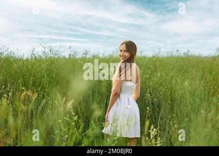 Joyeuse jeune femme en robe blanche courte dans le champ tournant en arrière et regardant par-dessus l'épaule Banque D'Images