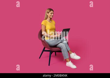 Femme blanche d'âge moyen souriante blonde dans une ambiance décontractée assise sur une chaise, dactylographiant sur un ordinateur portable Banque D'Images