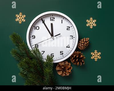 Horloge blanche et branche de sapin sur fond vert. Horloge de bureau comptant les dernières minutes avant la veille de Noël et le nouvel an. Concept vacances d'hiver. Banque D'Images