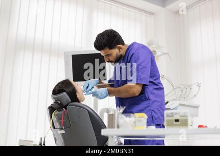 Un beau sourire aux dents blanches. Dentiste mâle barbu traitant les dents d'une jeune femme asiatique à l'aide d'un foret et d'une loupe dans le cabinet dentaire. Banque D'Images