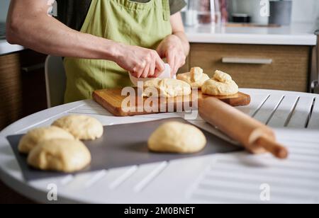 Boulanger de chef masculin méconnu en tablier vert faisant des boules de pâte. Couper de la pâte sur une planche à découper à l'aide d'une spatule. Cuisine maison, cuisine maison. Image de haute qualité Banque D'Images