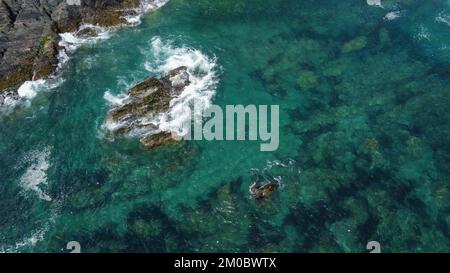 Rochers couverts de mousse noire au milieu des vagues turquoise de la mer celtique. Mousse de mer blanche sur les vagues. Eaux de l'océan Atlantique. Banque D'Images