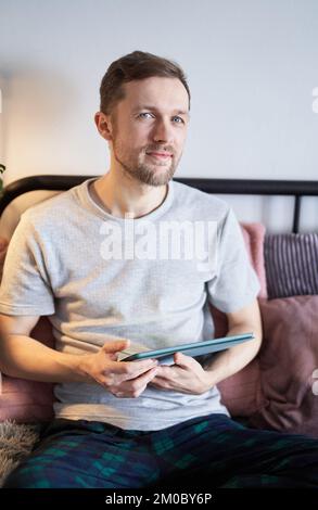 Homme joyeux à la maison travaillant avec une tablette. Jeune homme beau en t-shirt gris assis sur le lit faire des achats en ligne ou surfer sur Internet. Image verticale de haute qualité Banque D'Images