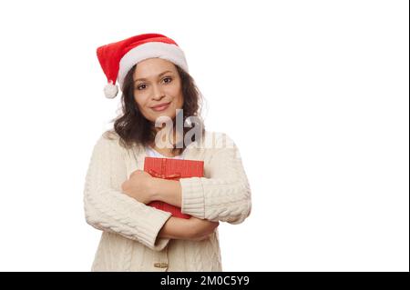 Jolie femme multi-ethnique portant un chapeau de père Noël et un chandail beige, avec boîte cadeau de Noël, sourires doux regardant l'appareil photo, isolée sur fond blanc Banque D'Images