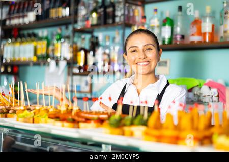 Le barman latino-féminin en uniforme sert de délicieuses tapas sur des brochettes au comptoir du bar Banque D'Images