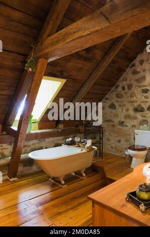 Salle de bains avec baignoire sur pied, mur de pierres de champ et panneaux de toit exposés à l'étage à l'intérieur d'une ancienne maison de style cottage Canadiana de 1722. Banque D'Images