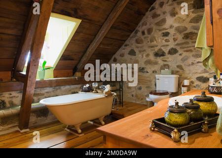 Salle de bains avec baignoire sur pied, mur de pierres de champ et panneaux de toit exposés à l'étage à l'intérieur d'une ancienne maison de style cottage Canadiana de 1722. Banque D'Images