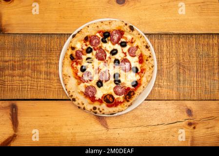 Une pizza napolitaine aux olives noires, au pepperoni, à la mozzarella, au parmesan râpé et à la tomate cuite dans un four en pierre sur une table en bois Banque D'Images