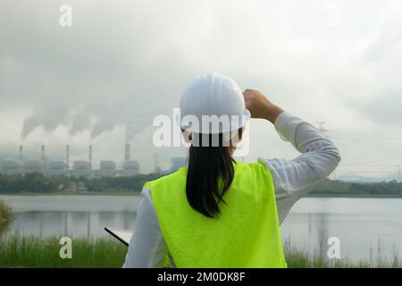 Une femme mécanicien en chef portant un gilet vert et un casque se tient à l'extérieur contre le fond de la centrale au charbon et de vapeur dans la brume matinale. Banque D'Images