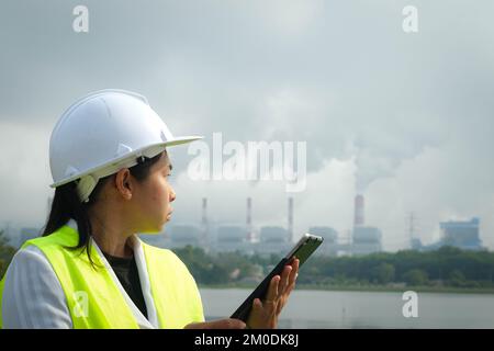 Une femme mécanicien en chef portant un gilet vert et un casque se tient à l'extérieur contre le fond de la centrale au charbon et de vapeur dans la brume matinale. Banque D'Images