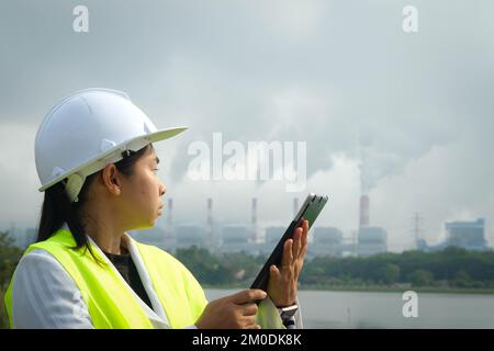 Une femme mécanicien en chef portant un gilet vert et un casque se tient à l'extérieur contre le fond de la centrale au charbon et de vapeur dans la brume matinale. Banque D'Images