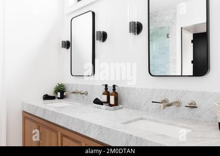 Une salle de bains confortable avec un meuble en bois, un comptoir en marbre et des sconces autour de miroirs carrés. Banque D'Images