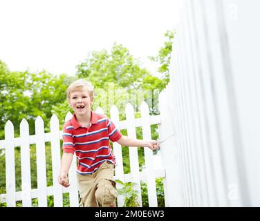 Oh pour être encore jeune. Un jeune garçon mignon courir un bâton le long d'une clôture de cornichon blanc dans son arrière-cour. Banque D'Images