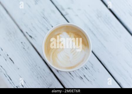 Cappuccino dans une tasse en papier sur une table en bois. Délicieux café du matin à emporter. Vue de dessus de la mousse un magnifique motif. Une tasse en papier jetable. Banque D'Images