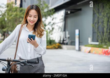 Portrait d'une femme d'affaires souriante qui fait le trajet entre son vélo et l'extérieur à l'aide d'un smartphone en milieu urbain Banque D'Images