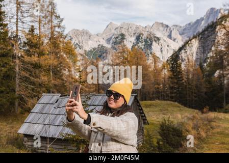 Portrait d'une jeune femme heureuse prenant un selfie devant des mélèzes dorés sous les montagnes en automne au col de Vrsic en Slovénie. Banque D'Images