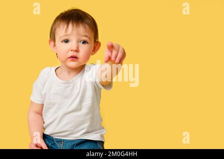 Portrait de bébé bouleversé sur fond jaune studio. Points enfant offensés vers l'avant avec index en t-shirt blanc et jeans bleus, copie de spac Banque D'Images