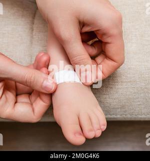 La mère colle un pansement médical adhésif sur la jambe du bébé. Main de maman avec ruban adhésif de protection collant et pied d'enfant. Enfant âgé de un vous Banque D'Images