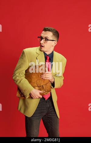 Portrait d'un jeune homme dans une veste élégante, des lunettes et un porte-documents vintage posé avec un visage sérieux sur fond rouge. Concept d'affaires, émotions Banque D'Images