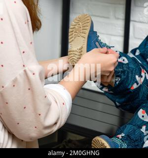 La mère met des chaussures bleues sur le pied de bébé enfant assis dans le couloir de la maison. Femme maman vêtue chaud bottes vêtements sur enfant pour l'hiver marcher dans c Banque D'Images