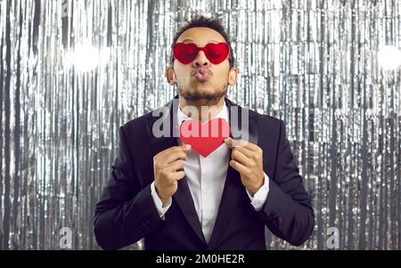 Drôle d'homme en costume et coeur verres tenant la carte de Saint-Valentin et de pouder ses lèvres pour baiser Banque D'Images