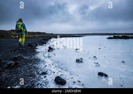 Islande, région de Sudurnes, Grindavik, péninsule de Reykjanes, la lagune bleue, un personnage portant une veste bleue et un sac à dos vert marche sur le bord d'une étendue d'eau chaude bleu turquoise résultant de l'activité géothermique naturelle Banque D'Images