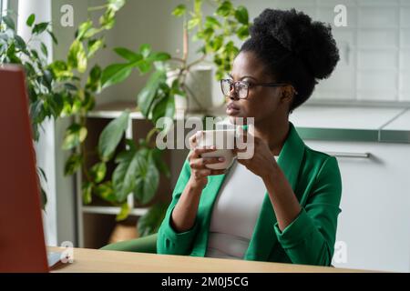 Une femme afro-américaine pensive qui a réussi à boire du café s'assoit à table avec un ordinateur au bureau à domicile Banque D'Images