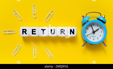 Concept de finance et d'économie. Sur fond jaune, un réveil bleu, des trombones et des cubes blancs sur lesquels le texte est écrit - RETOUR Banque D'Images