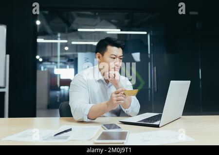 Achats en ligne, commande. Un jeune homme d'affaires asiatique est assis dans le bureau, regarde l'ordinateur portable et tient une carte de crédit dans sa main. Bien pensé, il fait des achats, passe une commande, et s'installe. Banque D'Images