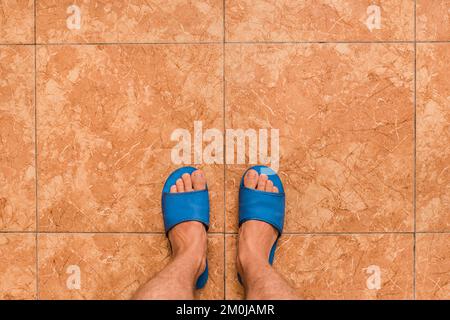 Pieds nus pour hommes, pantoufles bleues pour la maison, sur fond en céramique brun ou orange, vue d'en haut. Banque D'Images