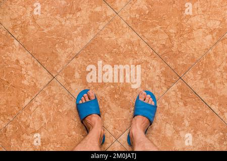 Pieds nus pour hommes, pantoufles bleues pour la maison, sur fond en céramique brun ou orange, vue d'en haut. Banque D'Images