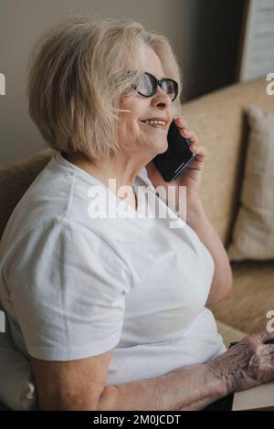 Femme âgée parlant avec un client sur un smartphone et écrivant dans un bloc-notes tout en étant assise sur un canapé à la maison. S'amuser à la maison, s'asseoir sur un canapé Banque D'Images