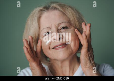 Femme blonde senior regardant la caméra, souriant appliquant des cellules oculaires transparentes isolées sur fond vert. Soins de beauté. Soin du visage Banque D'Images