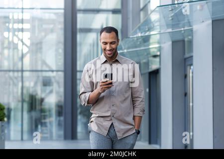 Homme d'affaires latino-américain dans une chemise décontractée à pied près du bâtiment de bureau de l'extérieur, homme souriant et en utilisant le téléphone, indépendant tapant le message et la navigation sur les pages Internet. Banque D'Images