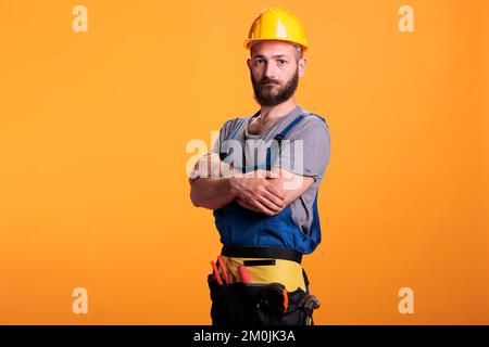 Charpentier réparateur gai avec uniforme rénovéant et casque de chantier posé sur la caméra en studio. Un homme de main professionnel se préparant à travailler sur un projet de rénovation, un expert artisanal. Banque D'Images