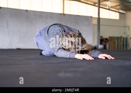 Une femme blonde de taille moyenne se reposant dans la pose de l'enfant pendant son entraînement de yoga de flux de Vinyasa seul et portant des vêtements de sport dans une salle de sport. Banque D'Images