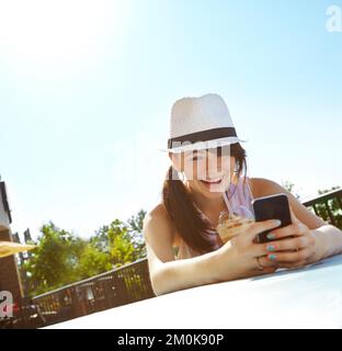 Soleil, smoothies et réseaux sociaux. Une bonne adolescente envoie un SMS sur un smartphone tout en dégustant un smoothie dans un café en plein air. Banque D'Images