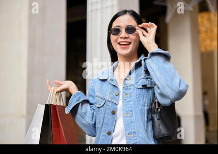 Belle et belle jeune femme asiatique portant des lunettes de soleil, portant ses sacs de shopping, aime faire du shopping au centre commercial de la ville. Banque D'Images