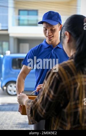 Un jeune livreur asiatique porte un uniforme bleu et remet le colis au client et le signe au client sur la tablette Banque D'Images