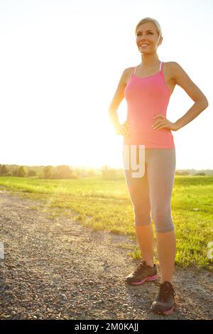 Profitez du paysage tout en vous relaxant. Une jeune femme en vêtement de sport debout dans une route de terre à côté d'un champ. Banque D'Images