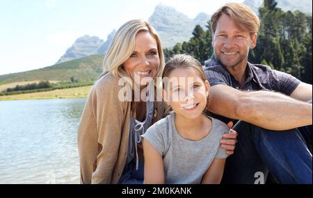 Des souvenirs qui durent toute une vie. Portrait d'une famille heureuse de trois personnes assises ensemble au bord d'un lac. Banque D'Images