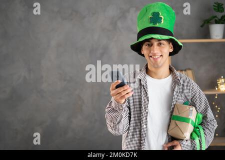 Homme arabe en lepretchun chapeau avec boîte cadeau de Saint Patrick sur smartphone et envoi de voeux de vacances, en regardant l'appareil photo. Gros plan portrait o Banque D'Images