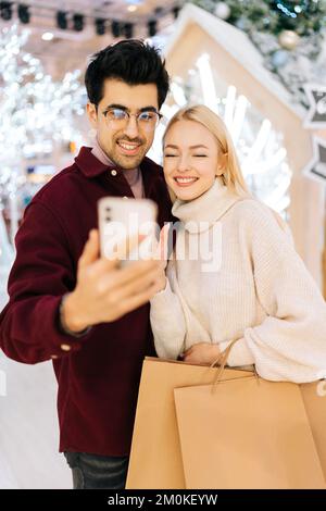 Photo verticale d'un couple heureux en train de parler en regardant un téléphone intelligent passer un appel vidéo, en donnant des salutations en ligne du nouvel an dans le hall des fêtes de shopping Banque D'Images