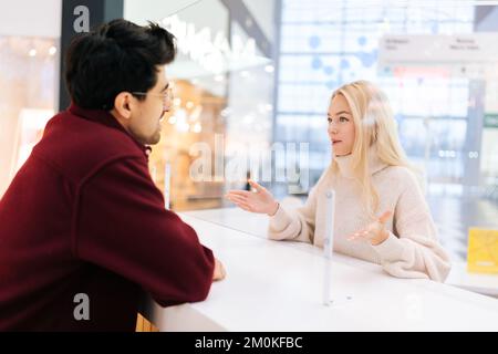 Vue arrière de l'épaule d'un homme souriant qui parle à travers une cloison en verre avec une femme blonde confuse debout à un bureau dans le hall lumineux du centre commercial. Banque D'Images