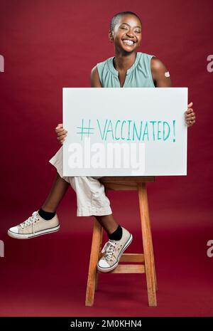 Femme vaccinée Covid afro-américaine montrant et tenant une affiche. Pleine longueur de femme noire souriante isolée contre arrière-plan rouge studio avec Banque D'Images