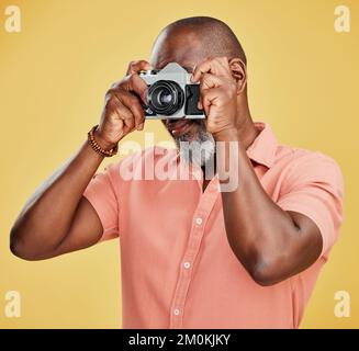 Un homme afro-américain debout seul sur fond jaune dans un studio et prenant des photos sur un appareil photo. Homme noir confiant tenant un appareil photo Banque D'Images