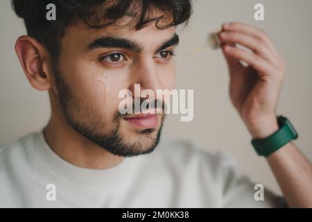 Homme appliquant du sérum facial et de l'huile essentielle pour des soins de la peau luisants, sains et esthétiques sur fond orange studio. Concept bien-être. Banque D'Images