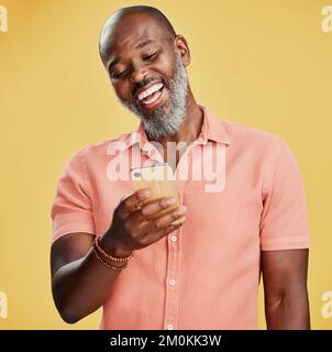 Un heureux Afro-américain homme debout contre un fond jaune de studio, tenant et utilisant son téléphone portable pour naviguer sur Internet. Homme noir souriant Banque D'Images